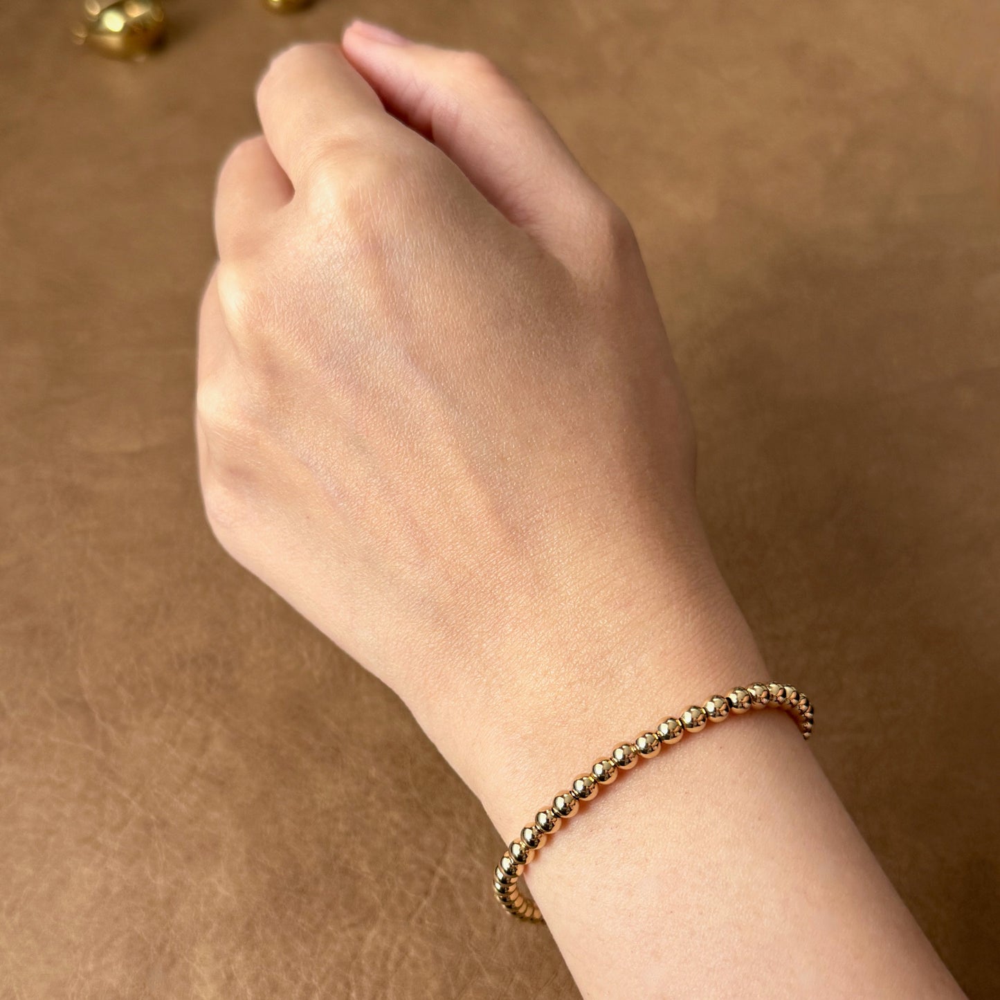 Hand wearing a gold bracelet on a brown background with gold beads.